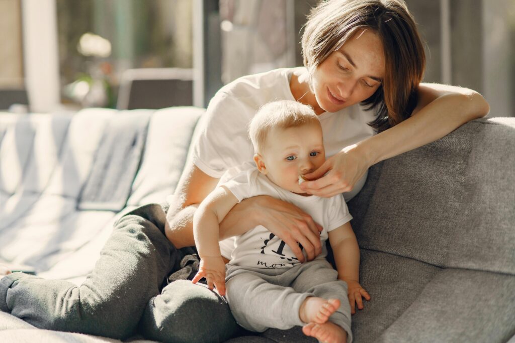 A mother lovingly relaxes with her baby on a sunny indoor sofa, exuding warmth and intimacy.