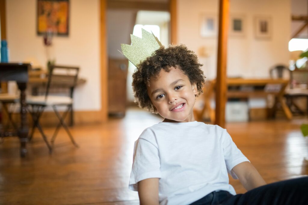 Smiling child with curly hair wearing a paper crown, sitting inside a warm home setting.
