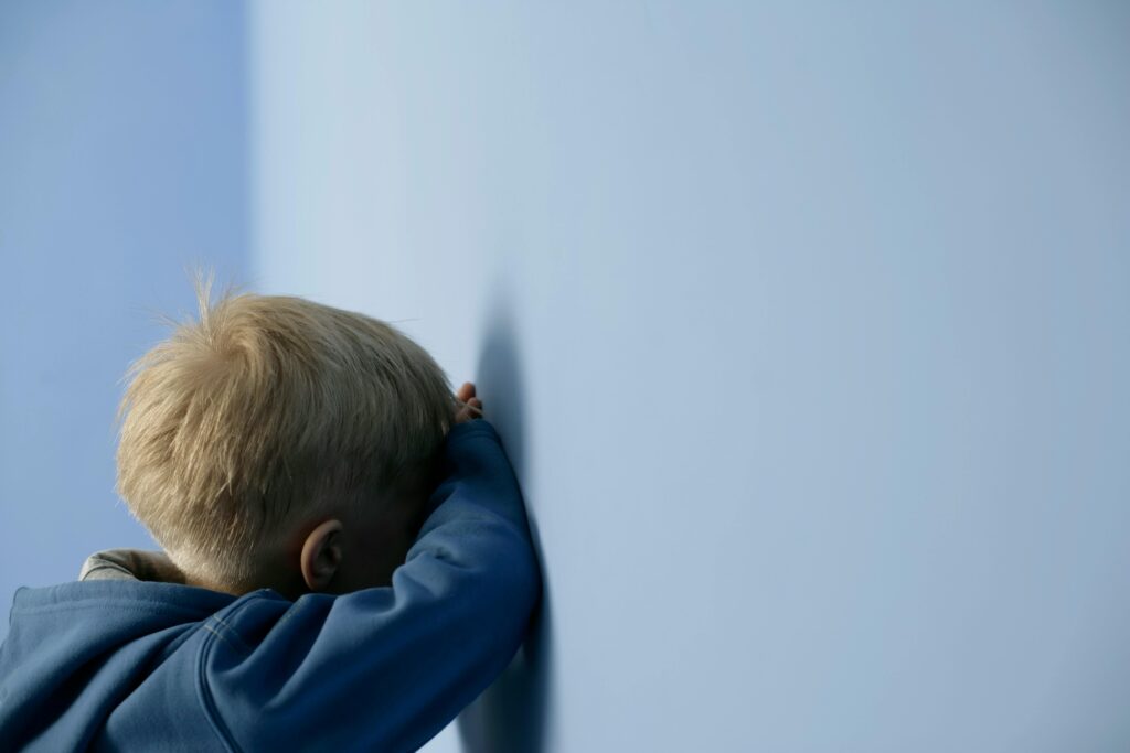 A young blonde child in a blue sweater leans against a blue wall, deep in thought.
