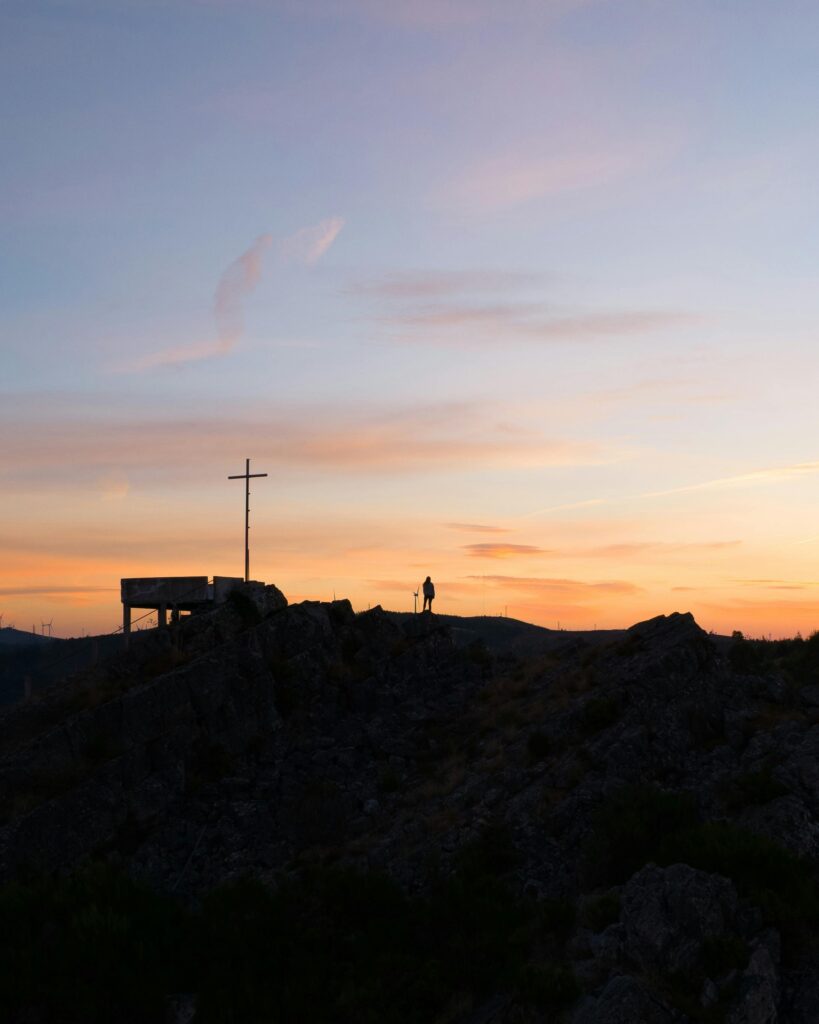 A lone figure stands by a cross silhouette against a vibrant sunset in Lousã, Portugal.
