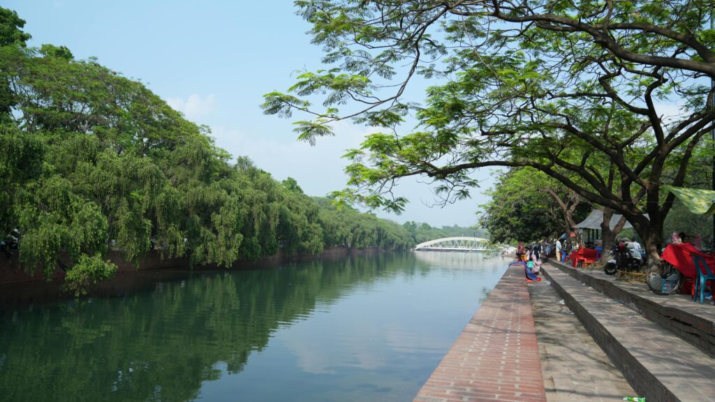 Scenic riverside park scene with trees, walkway, and a distant bridge in daylight.