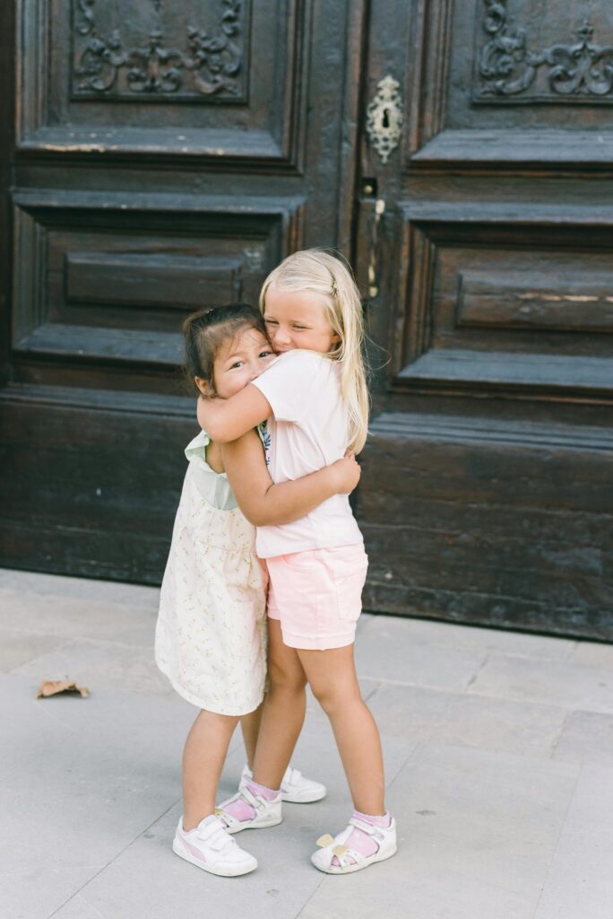 Two young girls share a heartfelt hug in front of a rustic wooden door, showcasing innocence and friendship.