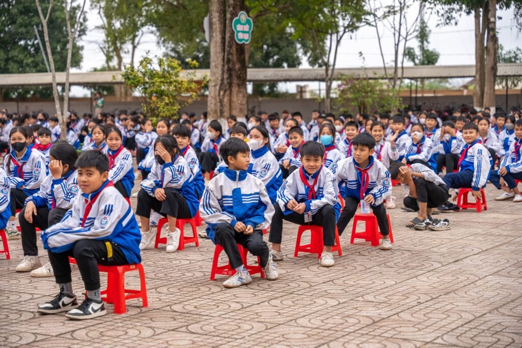 Students in uniform gathered for an outdoor school ceremony, sitting on red stools.