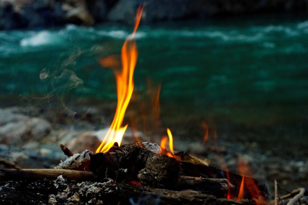 Close-up of a campfire with flames and embers by a flowing river, creating a serene outdoor scene.