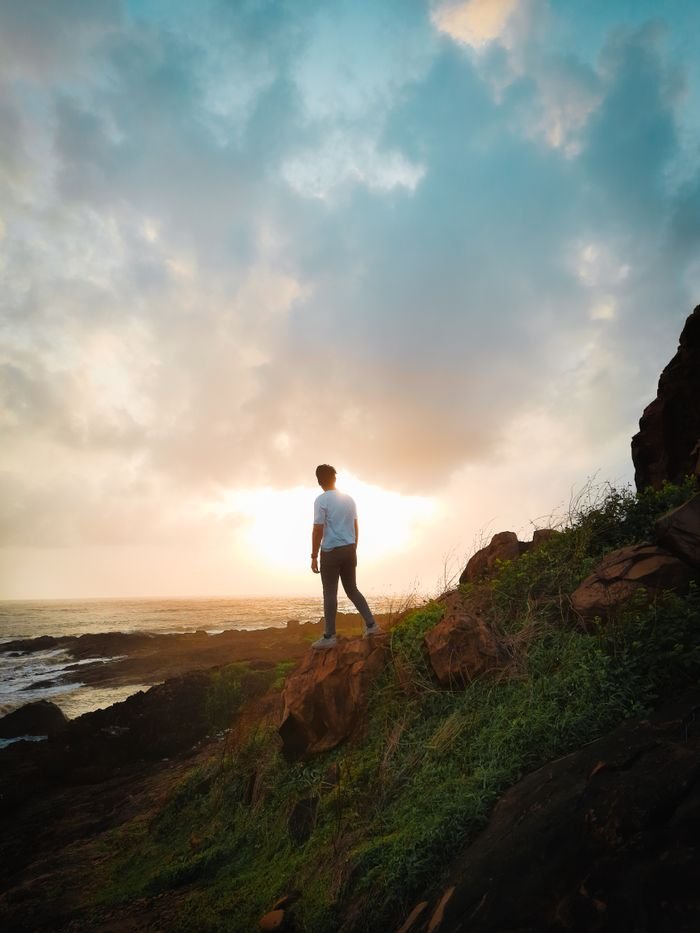 A lone person stands on a rocky coastal cliff, gazing at a stunning sunset over the ocean.